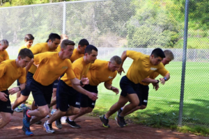 Action shot of a large group of midshipmen running on a dirt track near a fenced grass field