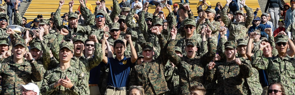 Large group of midshipmen in camouflage Navy working uniform cheering from the stands
