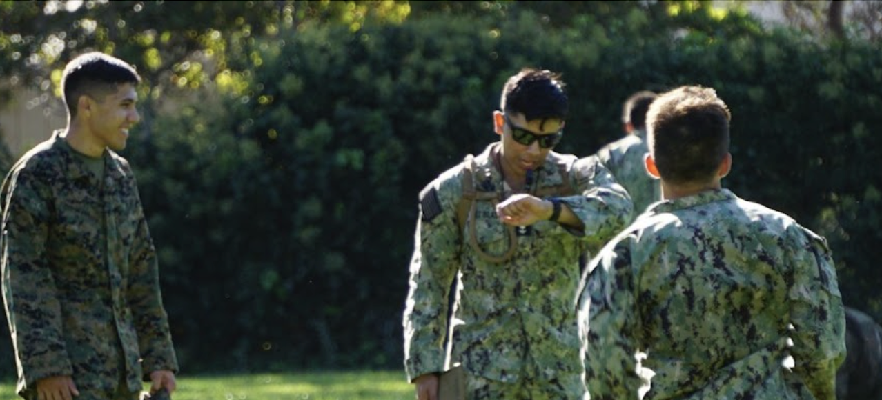 One of three midshipmen on campus at the green grassy North Field in camouflage uniform checking the time on wrist watch