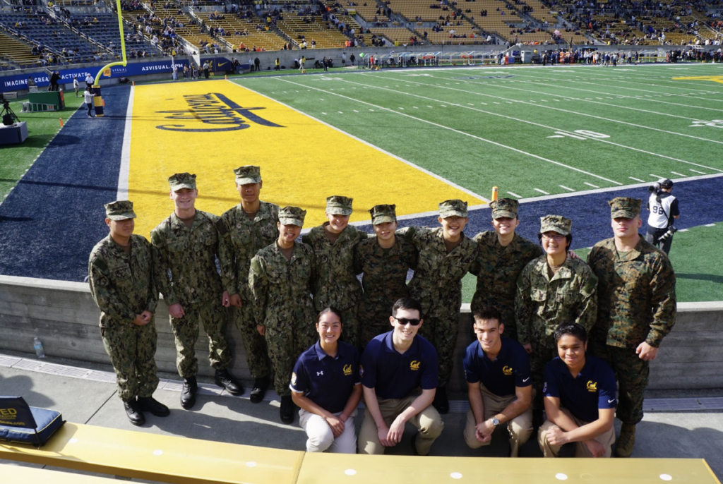 A group of fourteen midshipmen dressed in camouflage and polo uniforms on the bleachers and the California Memorial Stadium